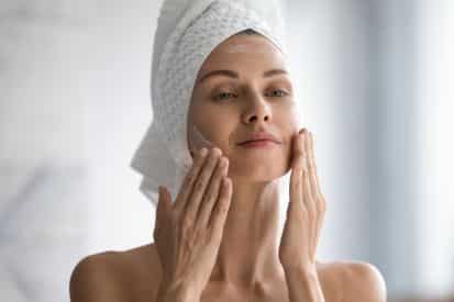 woman putting on face cream in the mirror with towel wrap on head